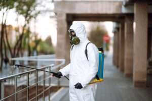 Man In Protective Suit And Mask Sprays Disinfector Onto The Railing In The Public Place. Covid 19.